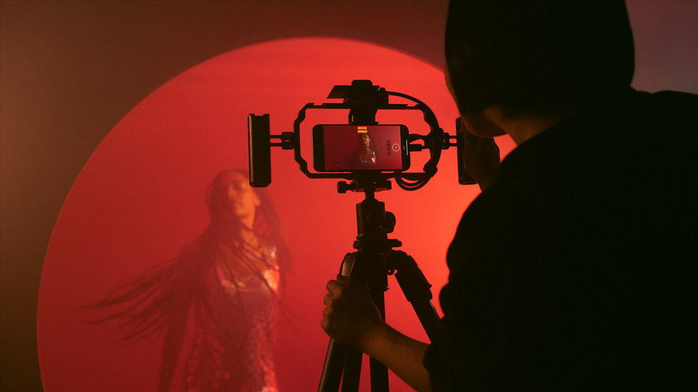 Una foto di una persona che usa un iPhone montato su un treppiede per filmare una donna su uno sfondo colorato