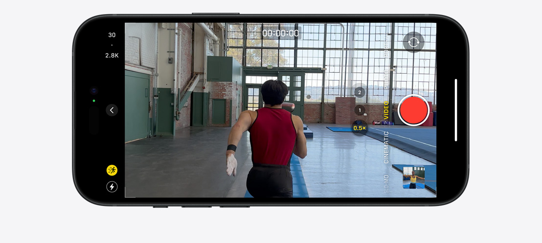 Obrázok iPhonu 15 Pro, ktorý v akčnom režime natáča gymnastu bežiaceho po veľkej osvetlenej miestnosti.