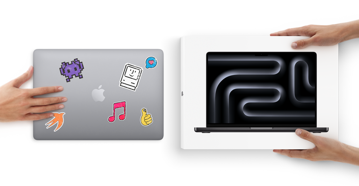 A closed silver Mac laptop covered with stickers and a new MacBook Pro in space black color still in its box being handled by pairs of hands
