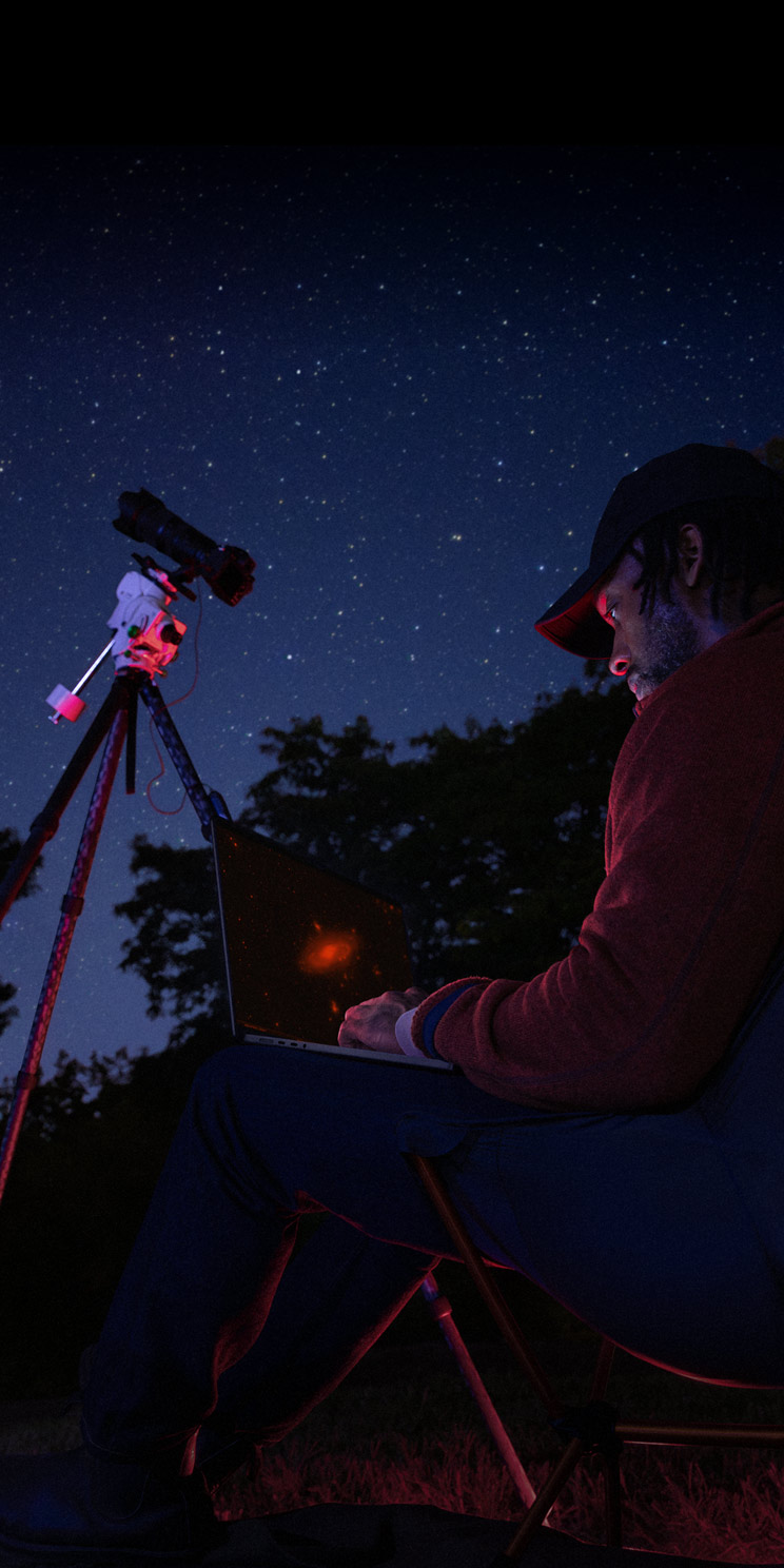 A person outside at night using a MacBook Pro on their lap, connected to a device on a tripod pointed toward the starry sky.