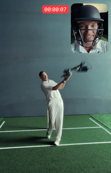 Cricketer Scott Boland stands in front of a wicket on an indoor cricket pitch holding a bat with iPhone 17 Pro attached, ready to strike. In the top right-hand corner, a picture-in-picture window shows a selfie view of cricketer Alex Carey wearing a helmet and talking.