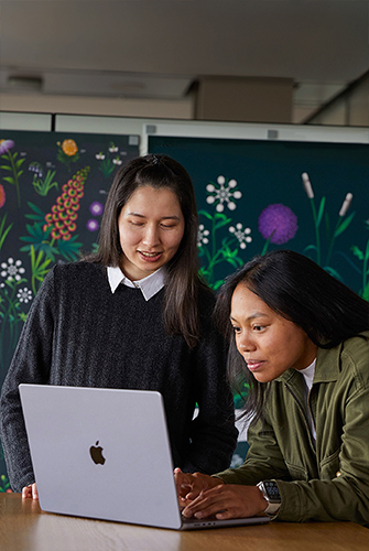 兩位 Apple 市場推廣團隊成員在其團隊設計的海報前，一起使用 MacBook 工作。