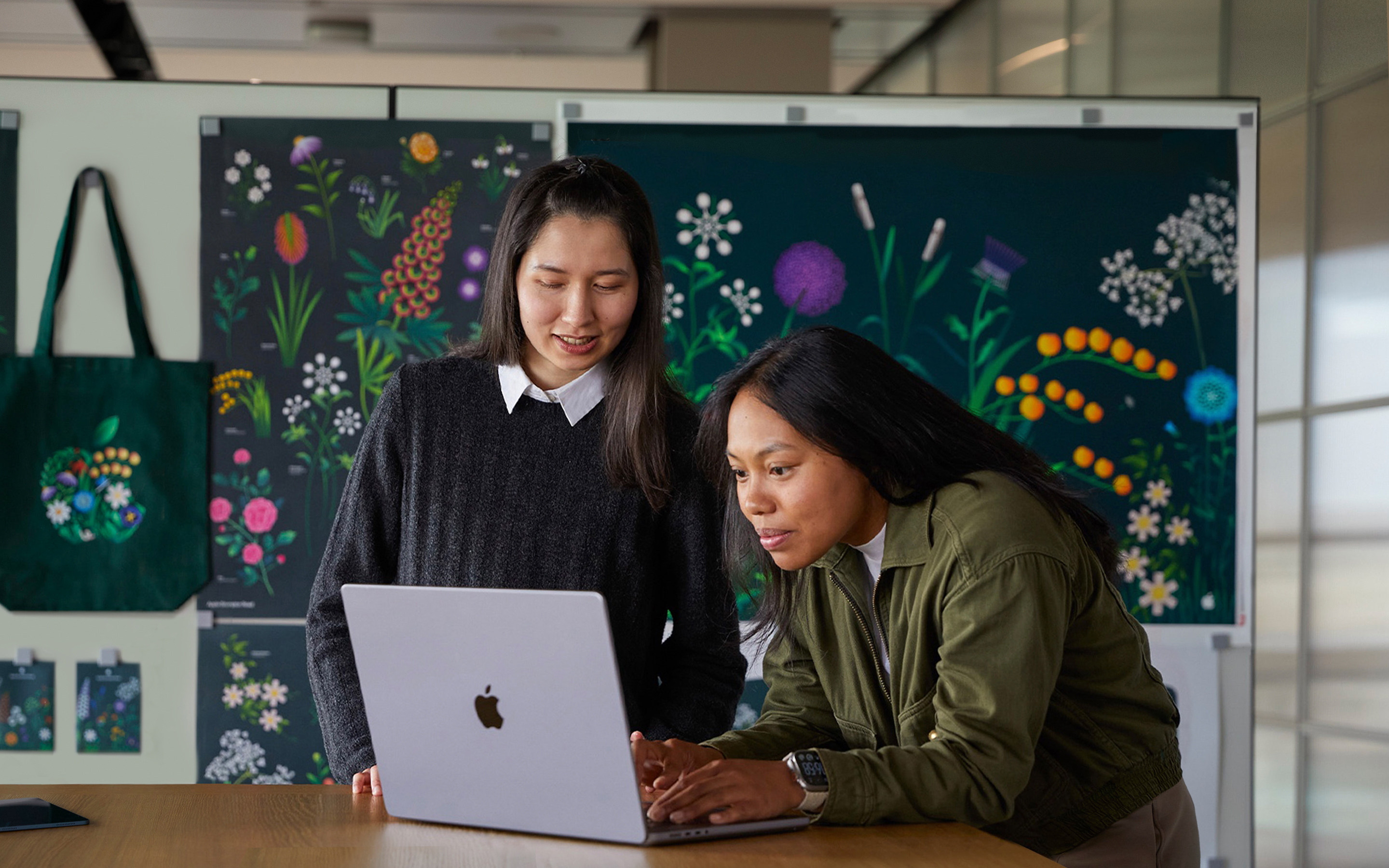 MacBook으로 작업 중인 두 명의 Apple 마케팅 팀원. 뒤쪽에는 마케팅 팀이 디자인한 포스터가 있음.