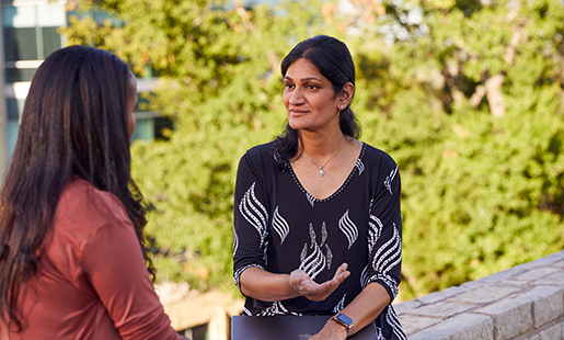 Ramani speaking with a colleague outside in an area landscaped with trees.