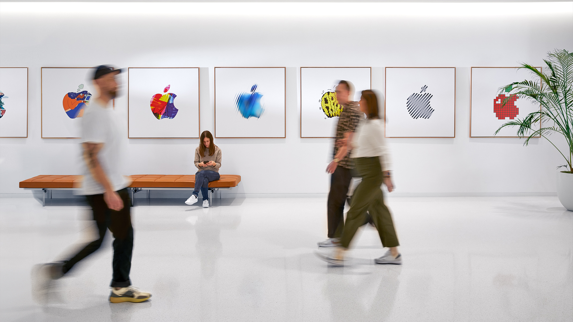 Three Apple colleagues walking past an interior wall with colourful Apple logos, and a fourth employee sitting on a bench.