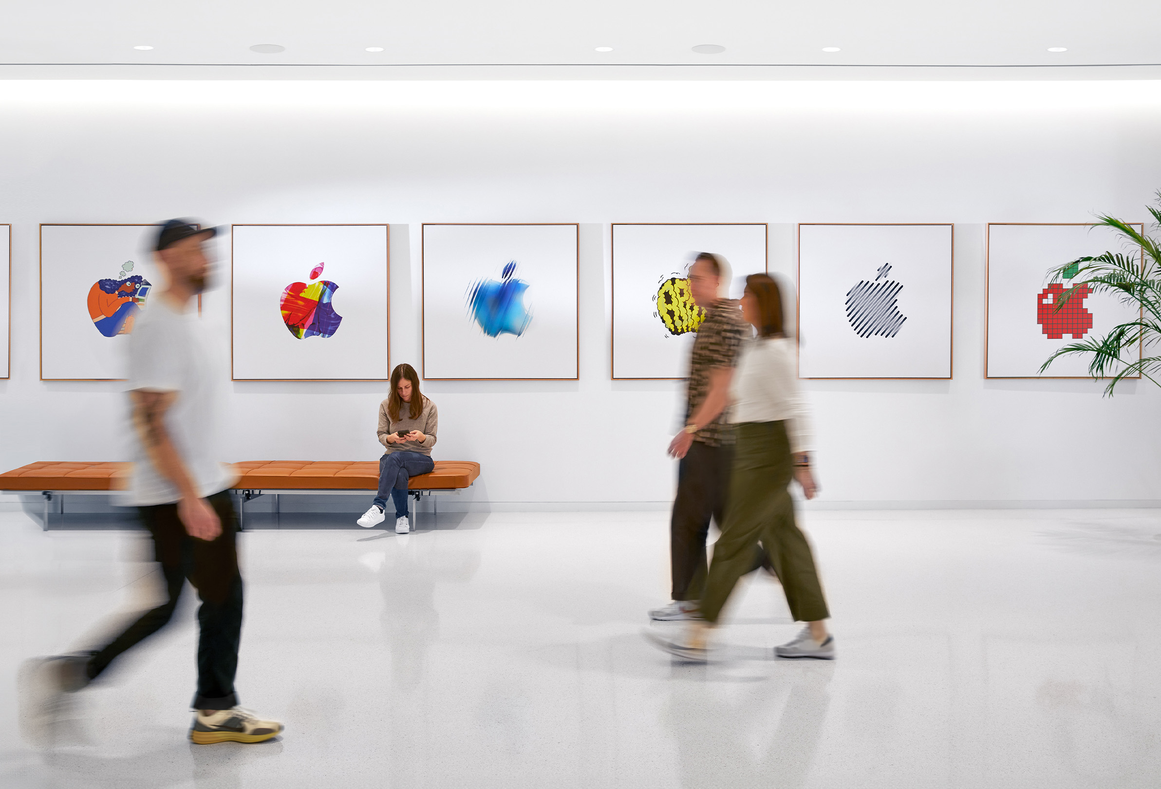 Three Apple colleagues walking past an interior wall with colourful Apple logos, and a fourth employee sitting on a bench.