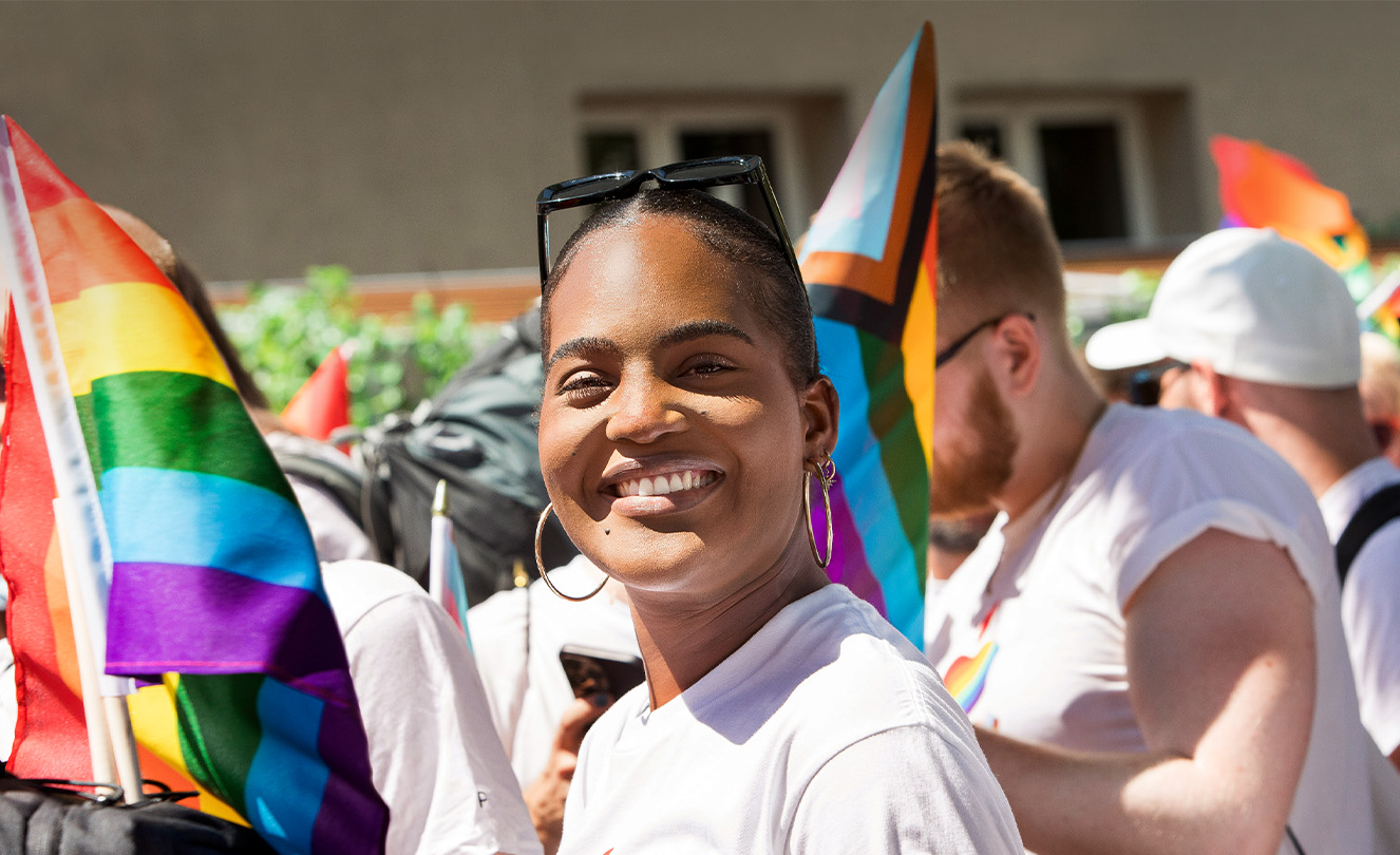 Uma funcionária da Apple sorridente num evento Apple Pride no meio de muitos funcionários que seguram bandeiras e usam t-shirts com as cores do arco-íris. 