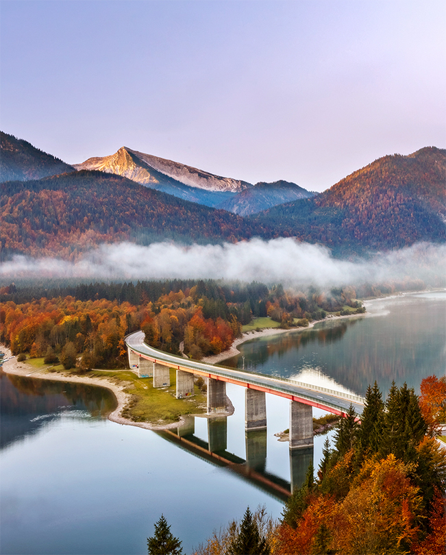 Imagem de uma ponte sobre um lago com montanhas ao fundo.