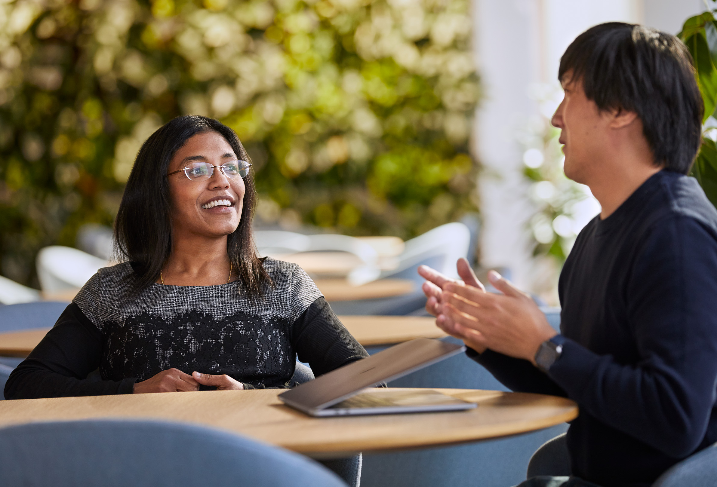 Dois funcionários da Apple sorriem sentados em uma área externa com um MacBook e folhagens ao fundo.