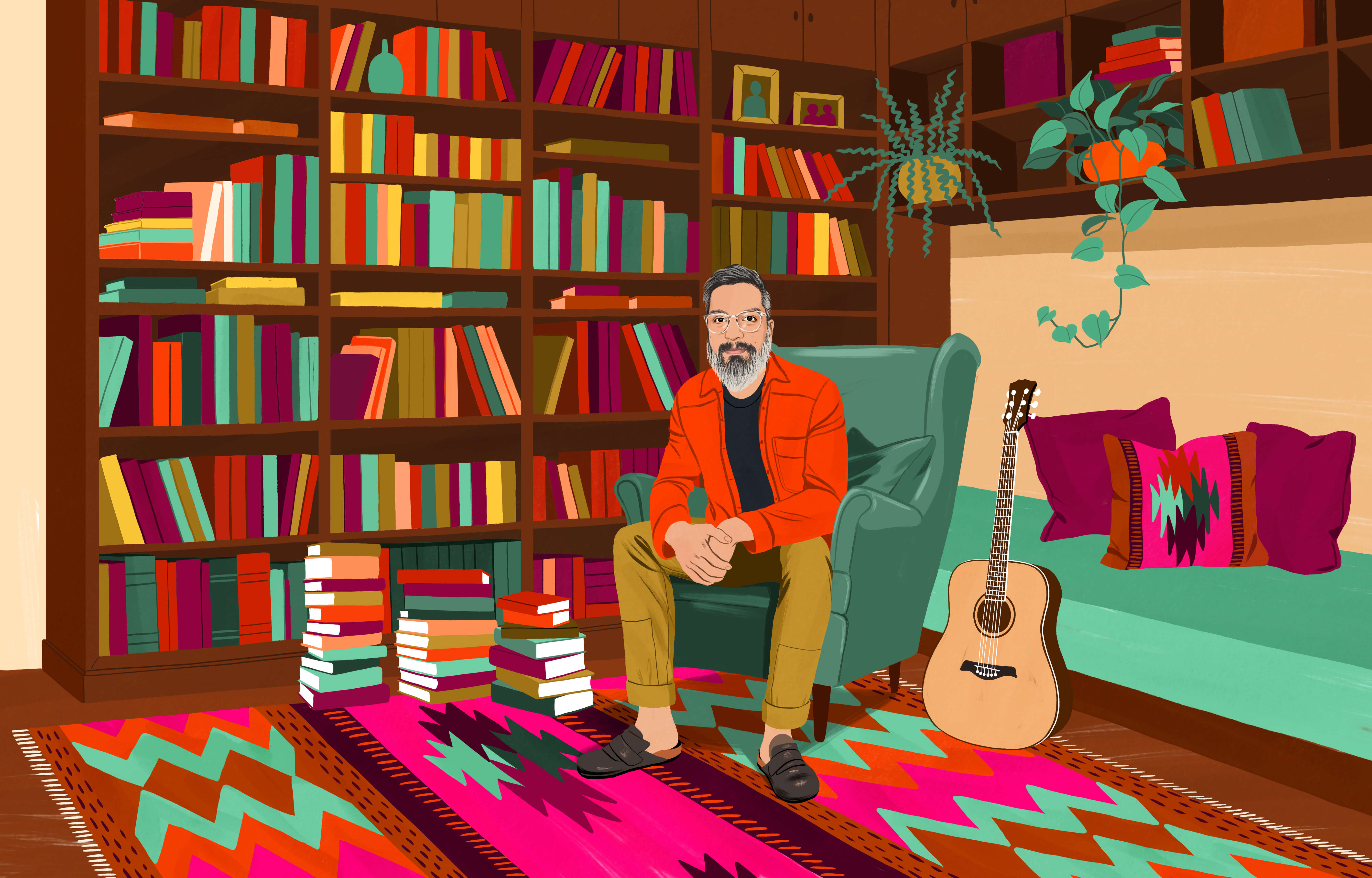 JP sitting in a wingback chair surrounded by many books in bookcases and stacked high on the floor. On the floor is a traditional Chilean rug with a colorful pattern. An acoustic guitar is within arm’s reach.