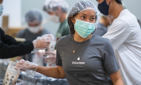 Une stagiaire d’Apple portant un t-shirt de bénévole Apple, sourit et regarde de côté tout en emballant des articles pendant un événement de bénévolat.