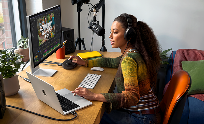 Žena sedí doma u pracovního stolu a používá MacBook Pro připojený k externímu monitoru, má nasazená sluchátka a mluví do mikrofonu