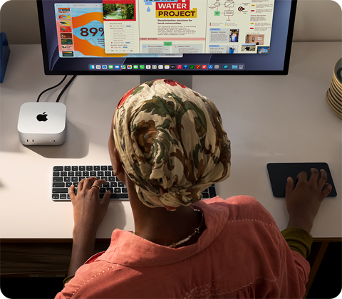 Inimene kirjutab laua taga, ees Mac mini, mis on ühendatud Studio Displayga, Magic Keyboard ja Magic Trackpad ning mitmesugused esemed