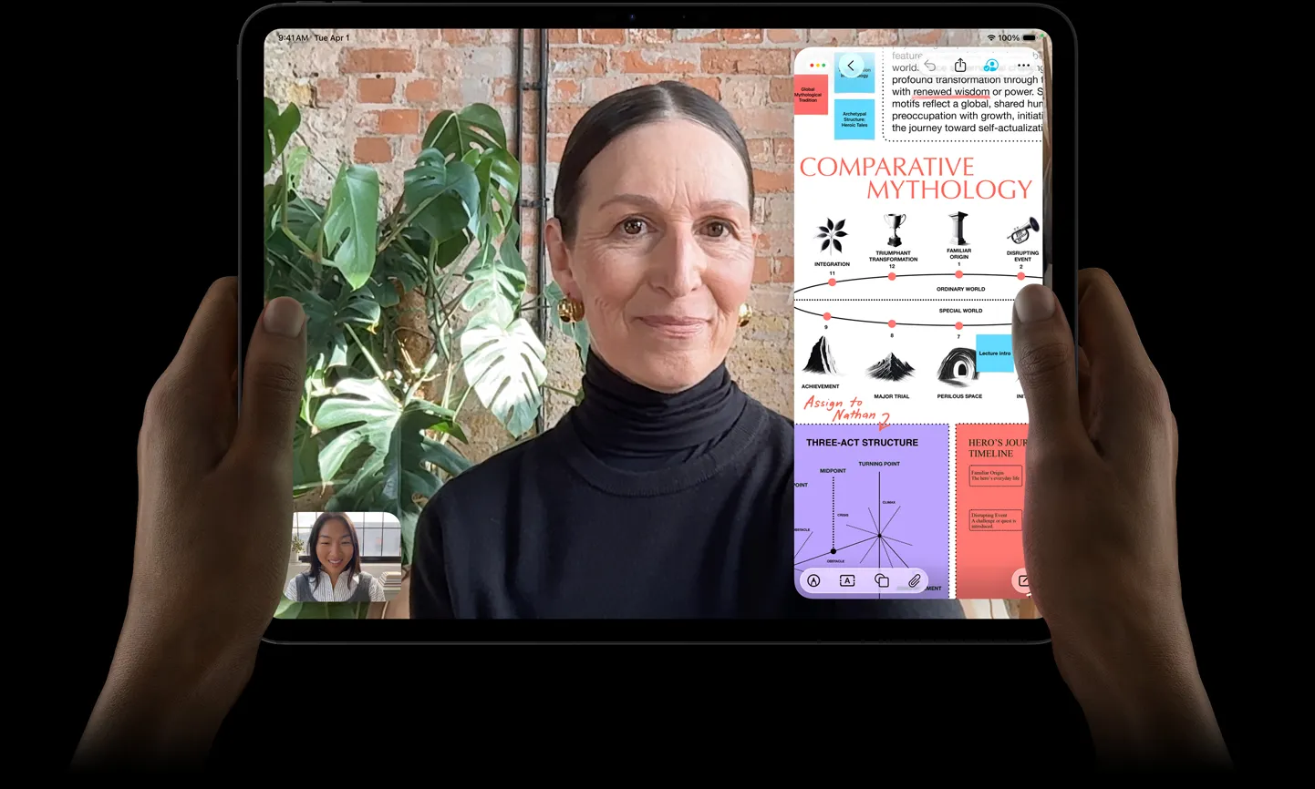 太空黑 iPad 機身正面，左右手以橫向握持，展示圓角設計，黑色顯示器邊框，顯示器顯示正在實時翻譯一段 FaceTime 對話，以及一個教育 app 展示「Comparative Mythology」簡報，其中有文字、圖表和手寫筆記