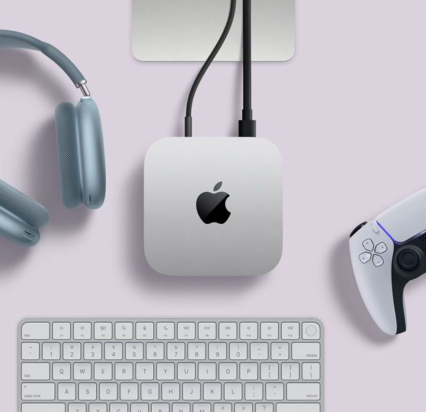 俯視圖展示銀色 Mac mini 擺放在桌上,有兩條連接線插在背面的連接埠,旁邊放有藍色 AirPods Max、一個銀色精妙鍵盤,以及一個無線遊戲手掣。