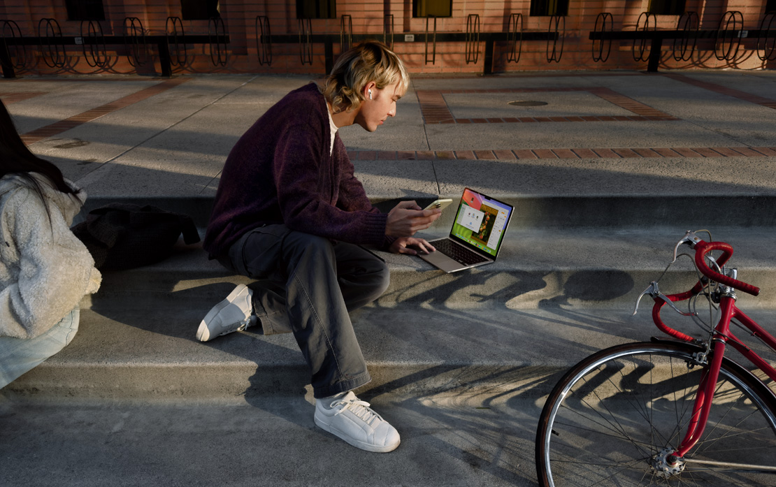 Seorang mahasiswa duduk di tangga di sebelah sepeda sambil menggunakan iPhone dan MacBook