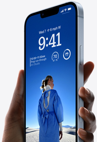 Tangan memegang iPhone 14 berwarna Biru dengan Layar Terkunci yang dipersonalisasi serta memperlihatkan foto seseorang dalam warna biru, waktu, dan widget.