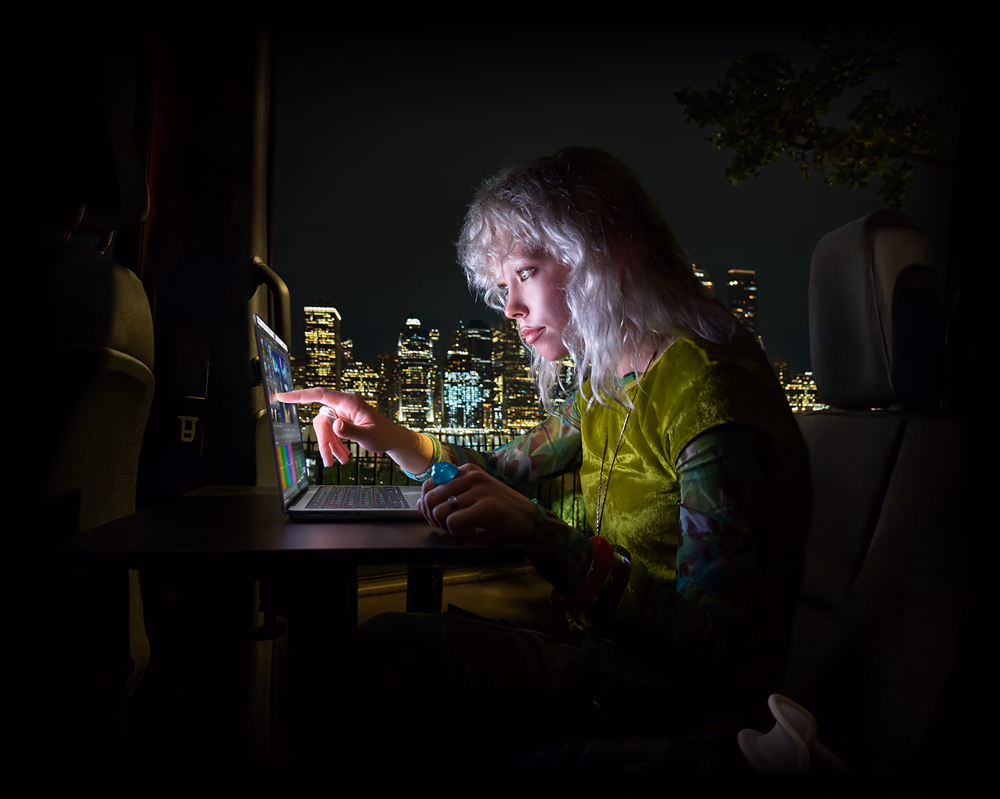 Seseorang duduk di kursi belakang mobil van, pintu penumpang yang terbuka memperlihatkan pemandangan kota pada malam hari, MacBook Pro dengan Layar PROLED XDR di atass meja kerja, cahaya dari layar menerangi orang tersebut  saat menggunakan fitur Sentuh untuk mengedit video di DaVinci Resolve Studio