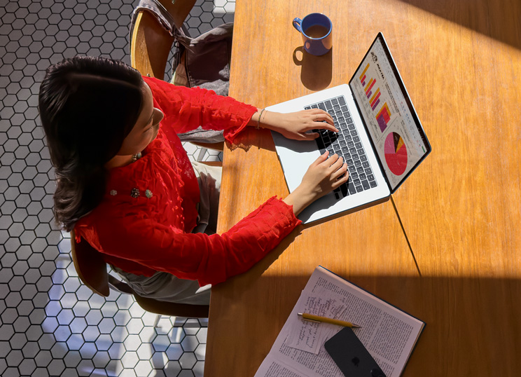 A person using Macbook at a table, with a mug, smartphone and papers nearby.