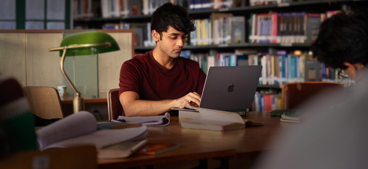 A person working on a MacBook at a library table, surrounded by open books and notes, with bookshelves in the background.