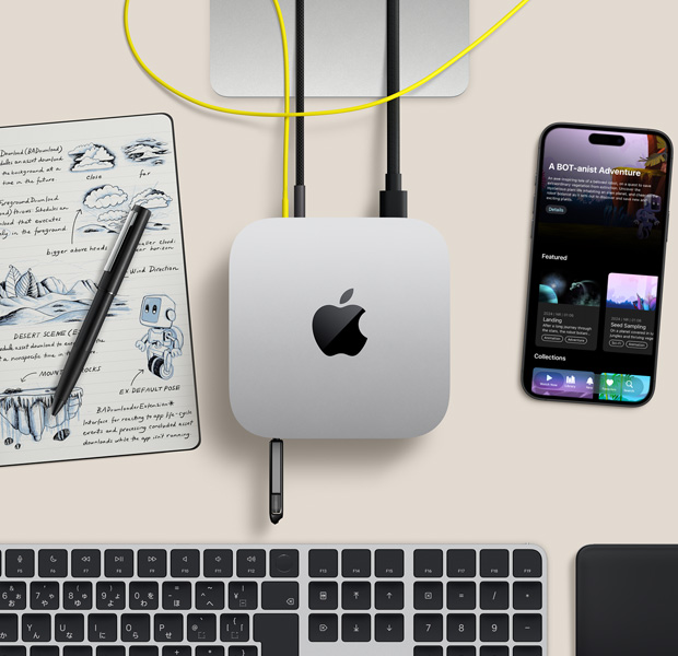 Mac miniを上から見た図。シルバーアルミニウム、丸みを帯びた四隅、中央に光沢のあるブラックのAppleのロゴ、コードで接続されている。手前にあるUSB-CポートにUSBメモリが挿入されている。Studio Displayのスタンドが一部見えている。iPhone、Magic Trackpad、Touch ID搭載Magic Keyboard、スタイラス、電子ペーパーのスケッチパッド