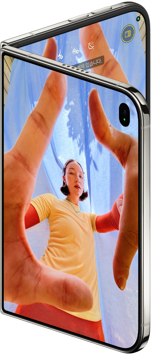 iPhone Duo 스타 화이트 색상, 살짝 열려 있는 상태. 외부 디스플레이에는 Center Stage 전면 카메라가 보이는 카메라 앱이, 내부 디스플레이에는 사진 앱 보관함이 표시되어 있습니다.