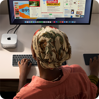 Žmogus rašo klaviatūra prie stalo, ant kurio matomi „Mac mini“, prie jo prijungtas „Studio Display“, „Magic Keyboard“, „Magic Trackpad“ ir įvairūs daiktai.