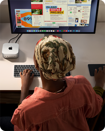 Persona pie rakstāmgalda raksta ar Mac mini, kas savienots ar Studio Display, Magic Keyboard tastatūru un Magic Trackpad skārienpaliktni, apkārt dažādi priekšmeti