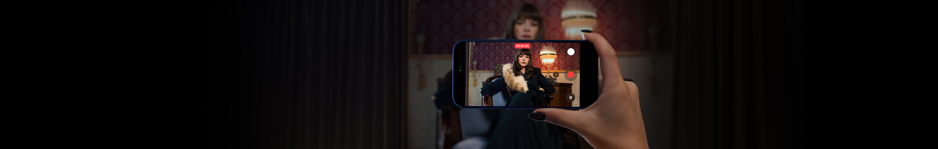 Mano sosteniendo un iPhone 17 Pro con la app de la cámara abierta, enfocando a Itatí Cantoral sentada en un sillón vestida de negro con cuello de piel.