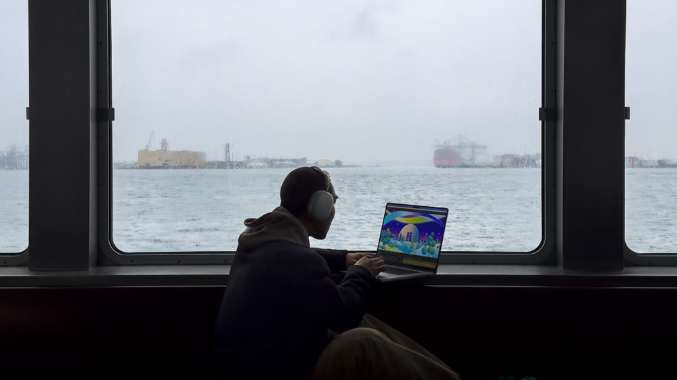 헤드폰을 착용한 사람이 창 밖으로 항구를 바라보며 새로운 MacBook Pro를 사용하는 모습.