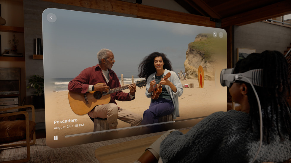 Apple Vision Proを着用して、リビングルームの窓に映し出された、海辺で音楽を演奏している人物2人の空間ビデオを観る人。