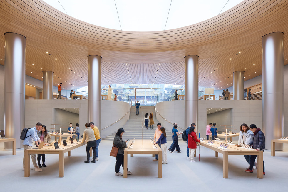 Clientes exploran una gama de productos Apple sobre mesas largas y usan la escalera central de la tienda.