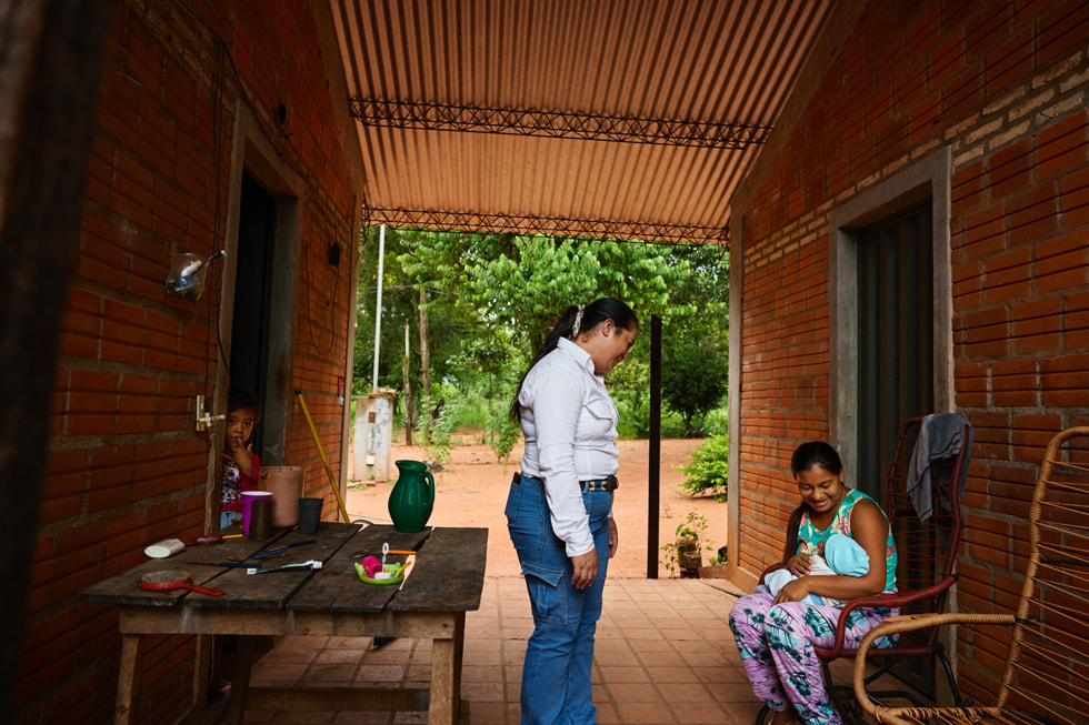 Gladys Nuñez står mellan två hus, vänd mot en person som håller en bebis i famnen medan ett barn tittar på från en dörröppning.