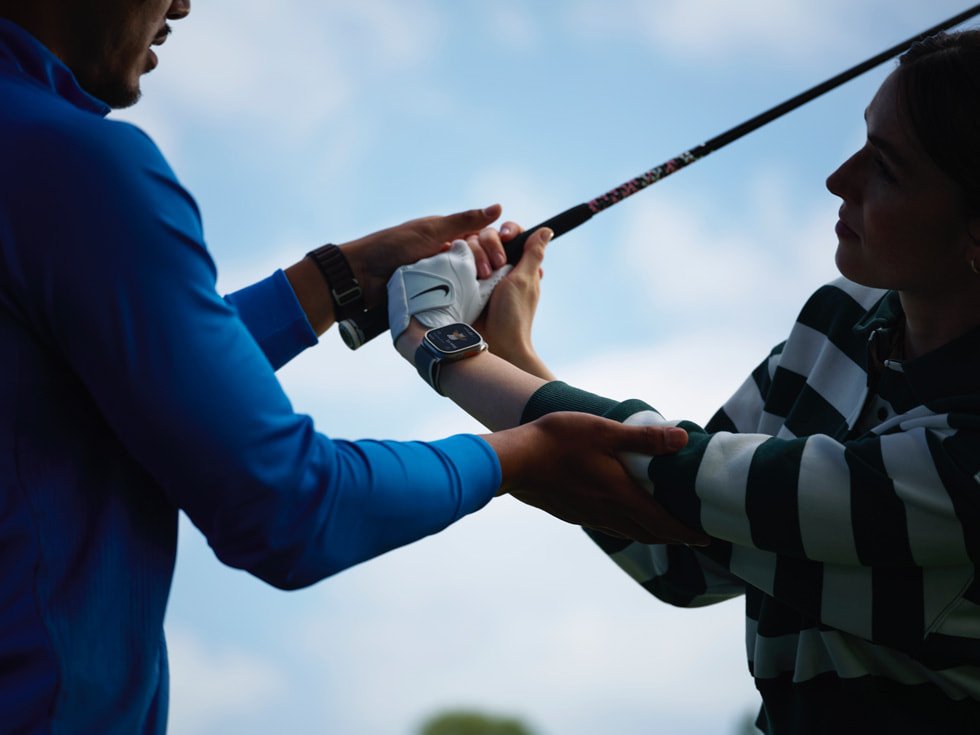 Foto pelatih golf membantu pegolf yang memakai Apple Watch sedang mengayunkan stik golf.