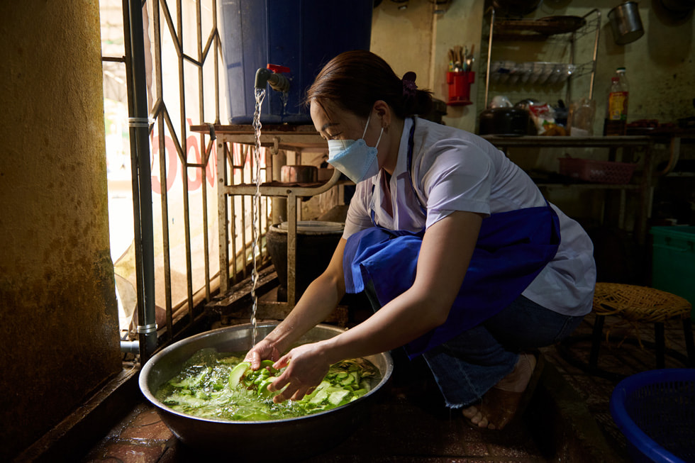 A person wearing a face mask and an apron washes vegetables in a giant basin with water pouring from a spigot.