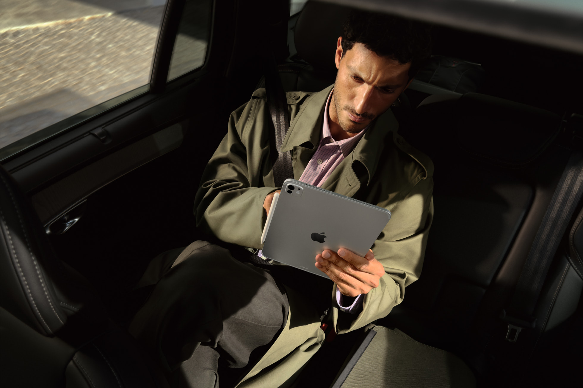 A person in a green trench coat sits in a car's back seat, focused on an iPad. Sunlight highlights their concentrated expression, evoking productivity.