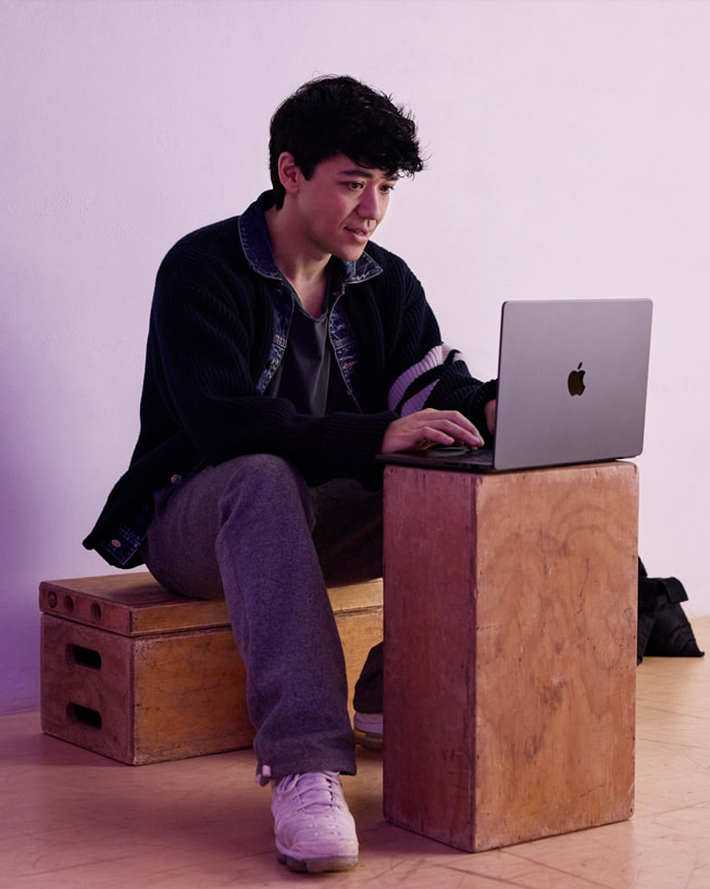 Kyle Hanagami ngồi trên một chiếc thùng gỗ trong không gian studio, gõ trên chiếc MacBook Pro được đặt trên một thùng khác.
