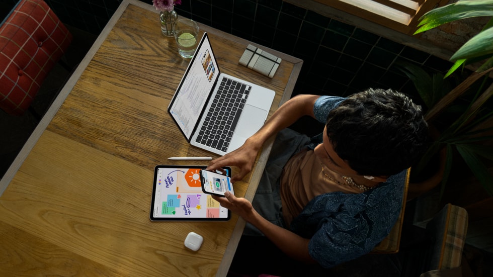 มุมมองด้านบนแสดงภาพบุคคลถือ iPhone และทำงานอยู่ที่โต๊ะพร้อม MacBook Pro ที่แสดงการเขียนโค้ดบนหน้าจอ พร้อมด้วย iPad, Apple Pencil และ AirPods