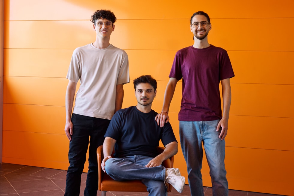 Nicolas Mariniello, Raffaele Fulgente e Fabio Pizzano, fondatori dello studio Flipping Hues, in piedi davanti a uno sfondo arancione.