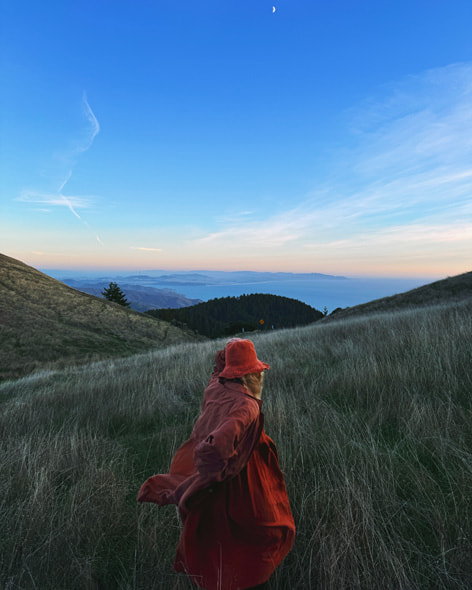 A photo of a person in red running through a mountainous field against a cloud-streaked blue sky with the Pacific Ocean in the background. Shot on iPhone 15 Pro Max by Anna-Alexia Basile.
