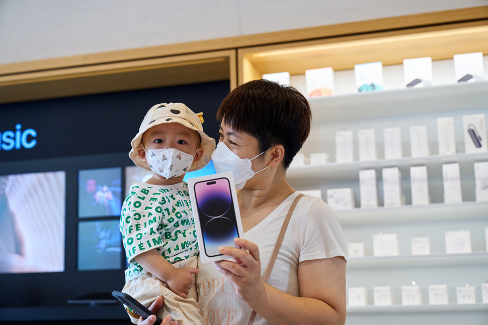 Apple 三里屯零售店內，一位顧客一邊手抱著孩子，一邊展示自己購買的 iPhone 14 Pro。