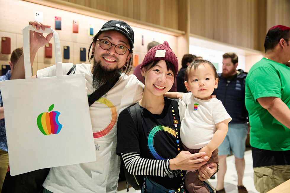 Two customers wearing Apple gear hold up an Apple tote while one carries a baby.