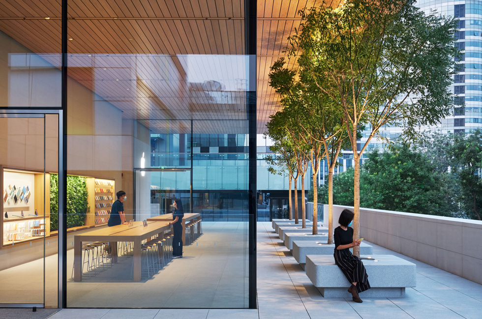 Een glazen hoek van de Apple Sanlitun en een zitgedeelte met bomen laten zien hoe naadloos de overgang van binnen naar buiten is.