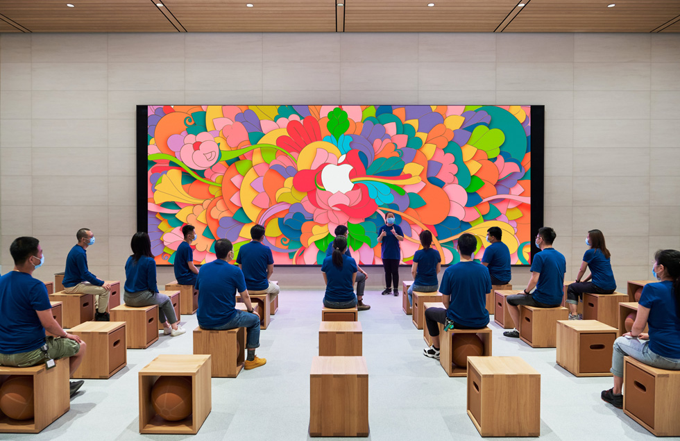 Apple employees, each wearing blue shirts and face masks and seated with social distancing in mind, listen to a presentation in the Forum. 