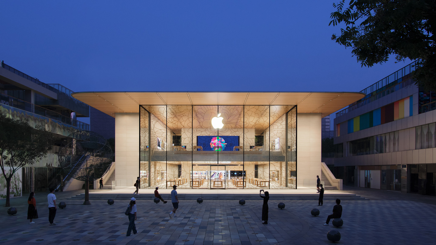 Buitenkant van de Apple Sanlitun in de avondschemer. Het verlichte interieur van de winkel benadrukt de transparantie en de flow van het ontwerp van het gebouw.