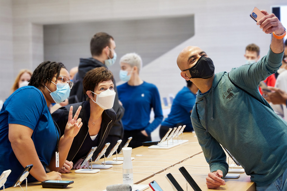 Un cliente del Apple Fifth Avenue se toma una selfie sobre una mesa de exhibición con Deidre O'Brien de Apple y un miembro del equipo.