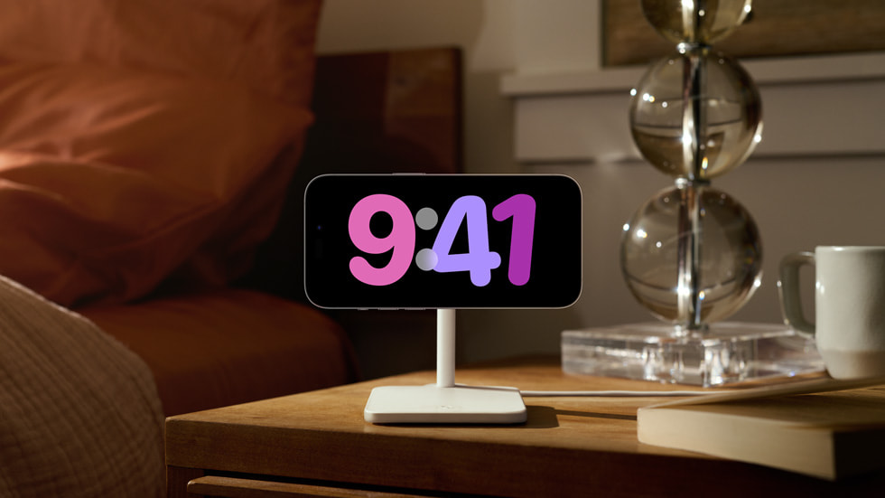 The full-screen StandBy experience is shown on an iPhone 14 Pro set on a user’s nightstand.