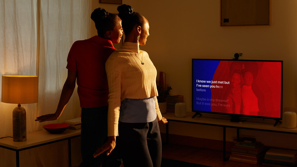 Two Apple users are shown posing back to back and mirrored on their TV screen.