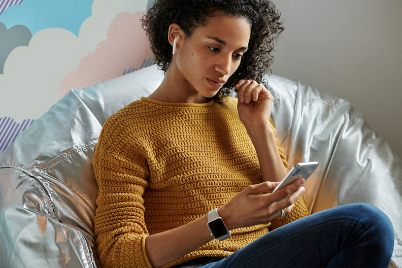 Mujer en un sofá sujetando un iPhone y escuchando música con los AirPods.