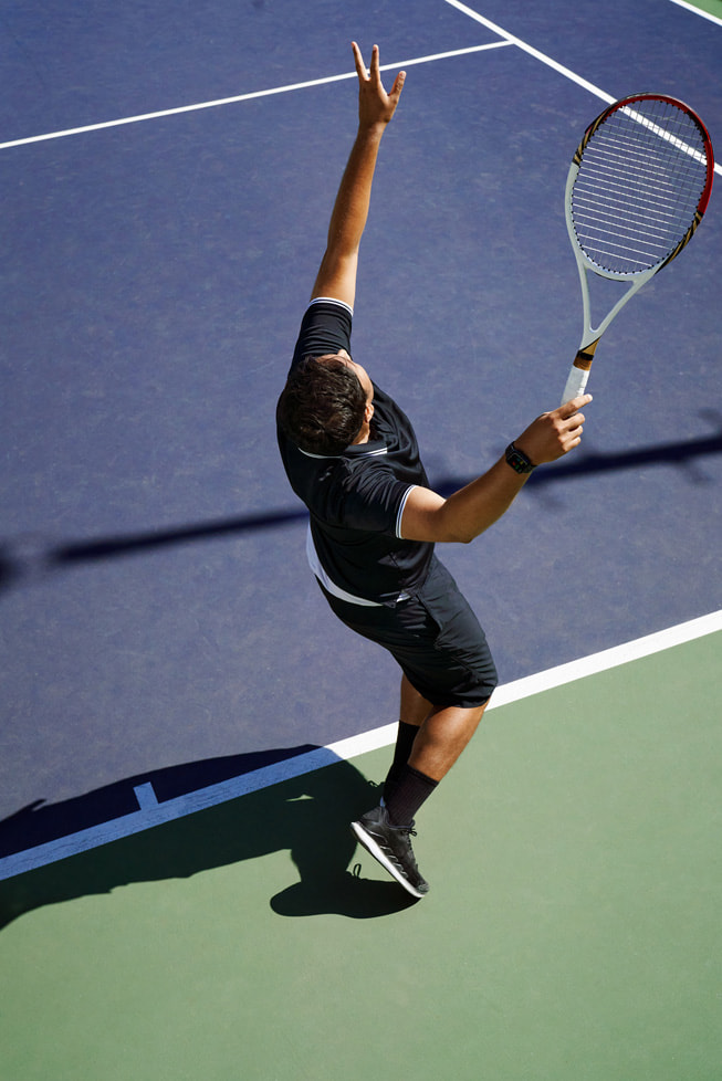 An overhead photo shows Swupnil Sahai, racquet in hand, reaching up in the midst of a serve on a tennis court.